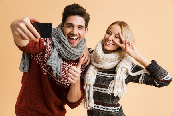 Portrait of a joyful young couple dressed in sweaters and scarves standing isolated over beige backgorund, taking a selfie together - Fotografie, Obrázek