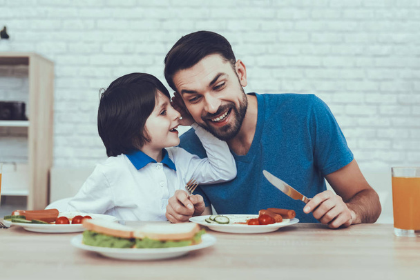 Man Spends Time with His Son. Father of Boy is Engaged in Raising Child. Father is Feeding a Son a Breakfast. Son Speaks in a Whisper to His Father. People is Sitting at Table. People on Kitchen. - Foto, Imagen