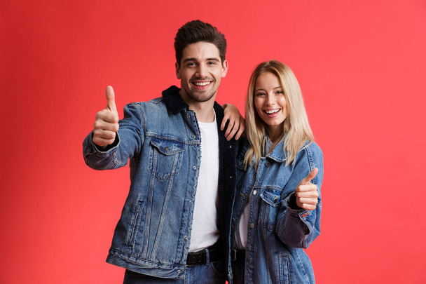 Портрет счастливой молодой пары, одетой в джинсовые куртки стоя вместе изолированы на красном фоне, показывая большие пальцы вверх
 - Фото, изображение