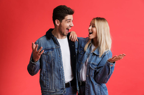 Portrait of an excited young couple dressed in denim jackets standing together isolated over red background, looking at each other - Φωτογραφία, εικόνα
