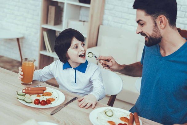 Man Spends Time with His Son. Father of Boy is Engaged in Raising Child. Father is Feeding a Son a Breakfast. Father is Giving a Cucumber to Son. People is Sitting at Table. People Located on Kitchen. - Fotoğraf, Görsel