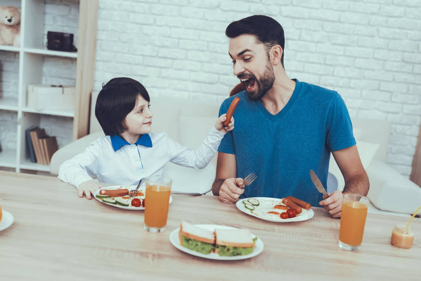 Man Spends Time with His Son. Father of Boy is Engaged in Raising Child. Father is Feeding a Son a Breakfast. Son is Giving a Sausage to Father. People is Sitting at Table. People Located on Kitchen. - Fotoğraf, Görsel