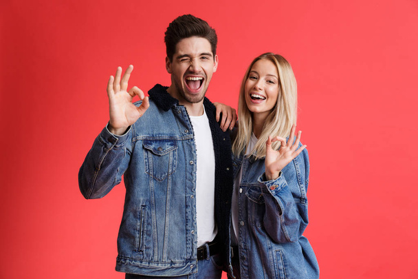 Portrait of a happy young couple dressed in denim jackets standing together isolated over red background, showing ok gesture - Foto, Imagem