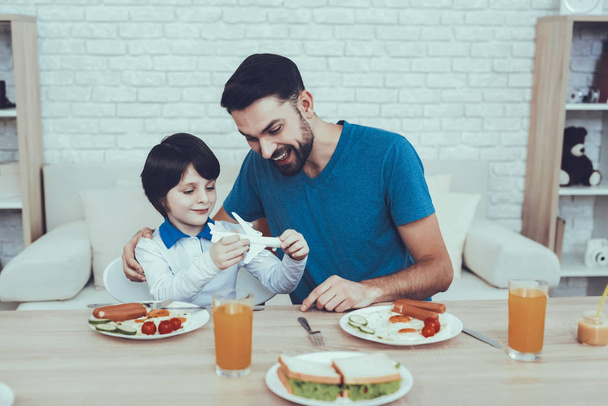 Man Spends Time with His Son. Father of Boy is Engaged in Raising Child. Father is Feeding a Son a Breakfast. Son is Holding a Toy Plane. People is Sitting at Table. People Located on Kitchen. - Φωτογραφία, εικόνα