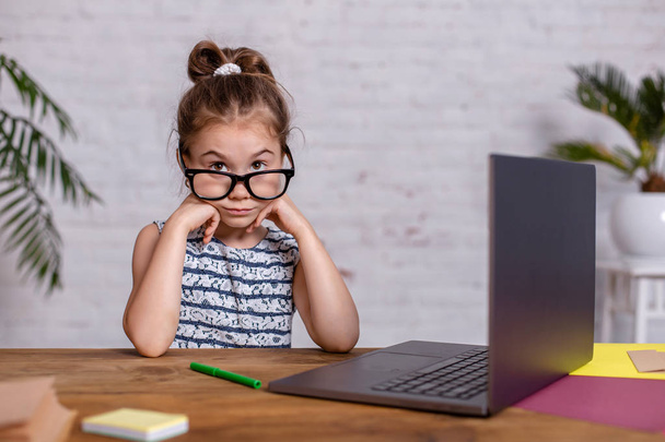 Petite fille mignonne est assise à table avec son ordinateur portable et son carnet, portant des lunettes
 - Photo, image