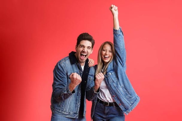 Portrait of a happy young couple dressed in denim jackets standing together isolated over red background, celebrating success - Foto, immagini