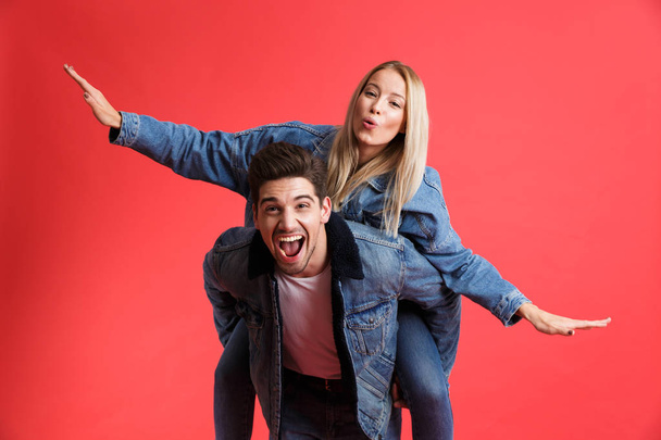 Portrait of a joyful young couple dressed in denim jackets standing together isolated over red background, piggyback ride - Foto, imagen