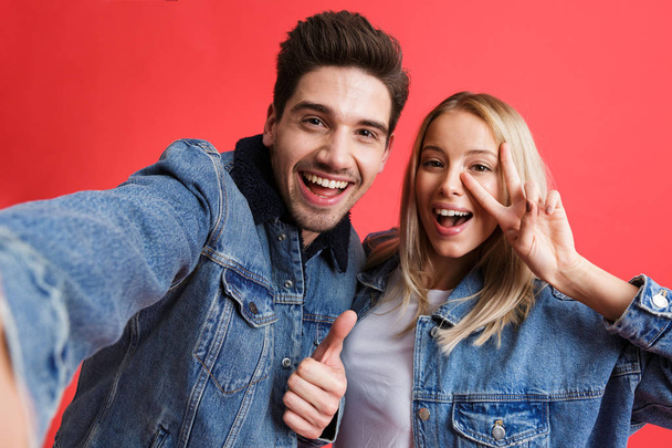 Portrait of a happy young couple dressed in denim jackets standing together isolated over red background, taking a selfie, giving thumbs up - Φωτογραφία, εικόνα