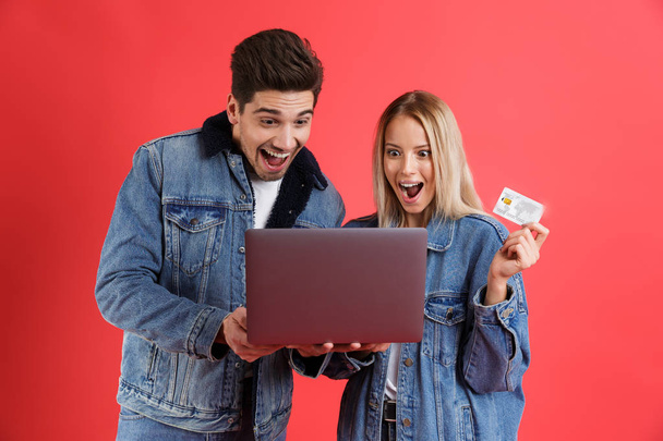 Portrait of an excited young couple dressed in denim jackets standing together isolated over red background, looking at laptop computer, holding plastic credit card - Foto, Bild
