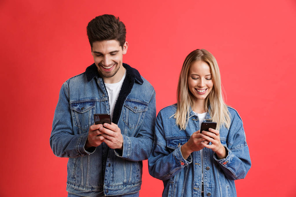 Portrait of a smiling young couple dressed in denim jackets standing together isolated over red background, using mobile phones - Foto, Bild
