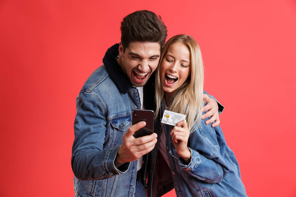 Portrait of a smiling young couple dressed in denim jackets standing together isolated over red background, using mobile phone, holding credit card, hugging - 写真・画像
