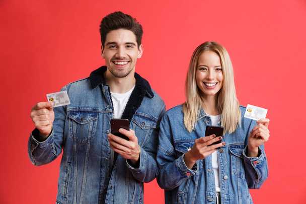 Image of excited young loving couple in jeans denim jackets using mobile phones isolated over red background holding credit cards. - 写真・画像