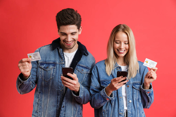 Portrait of a smiling young couple dressed in denim jackets standing together isolated over red background, using mobile phones, showing credit cards - 写真・画像