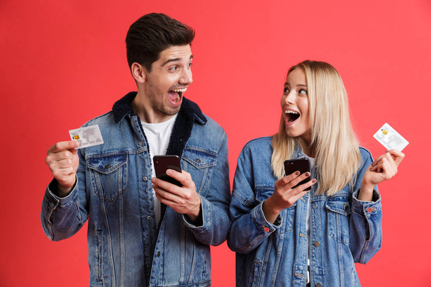 Portrait of an excited young couple dressed in denim jackets standing together isolated over red background, using mobile phones, showing credit cards, looking at each other - Foto, afbeelding