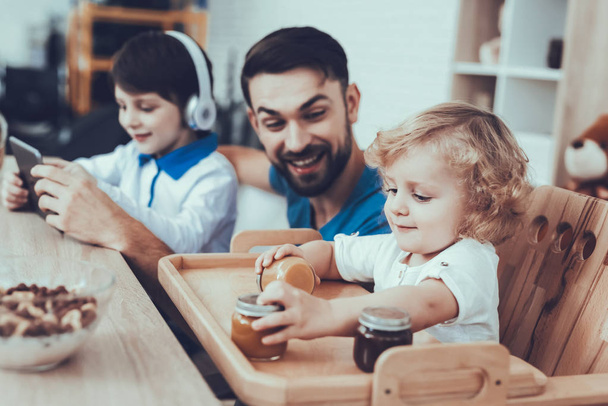 Man Spends Time with His Sons. Father is Engaged in Raising Children. Eldest Son is Watching Video on Tablet PC. Younger Son is Playing with Baby Food. Boys is Sitting at Table. People on Kitchen. - Foto, Imagem