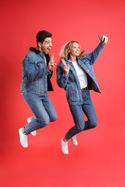 Full length portrait of a cheerful young couple dressed in denim jackets jumping together isolated over red background, taking a selfie - Фото, зображення