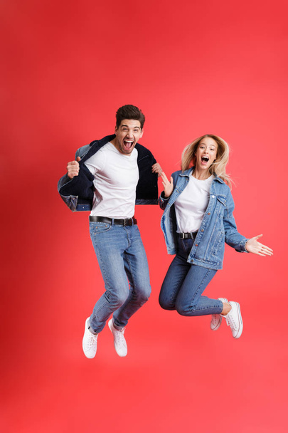 Full length portrait of a cheerful young couple dressed in denim jackets jumping together isolated over red background - Φωτογραφία, εικόνα