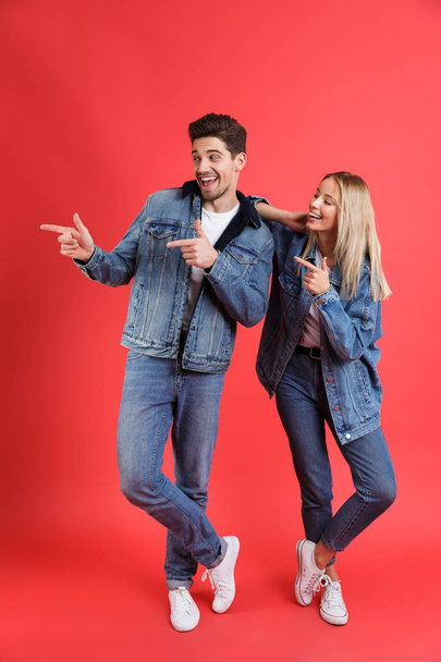 Full length portrait of a cheerful young couple dressed in denim jackets standing together isolated over red background, pointing fingers away at copy space - Zdjęcie, obraz