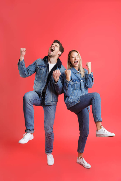 Full length portrait of a cheerful young couple dressed in denim clothes isolated over red background, celebrating success - Photo, Image