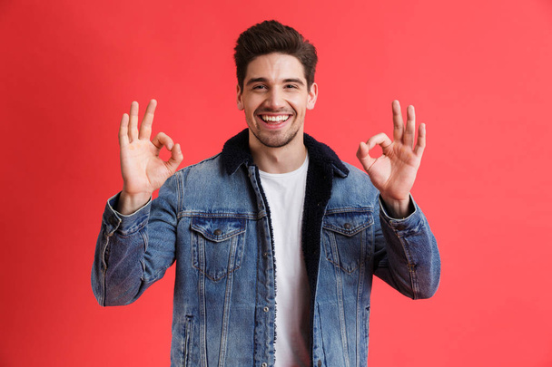 Portrait of a cheerful young man dressed in denim jacket standing isolated over red background, showing ok - Фото, зображення
