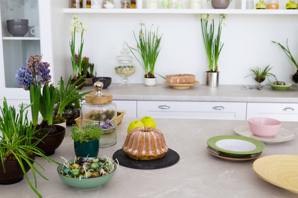 Μοντέρνα φωτεινή κουζίνα με τραπέζι, πράσινα λουλούδια, τούρτα, πλάκες, λευκά έπιπλα. Έννοια της εσωτερικό κουζινών. - Φωτογραφία, εικόνα