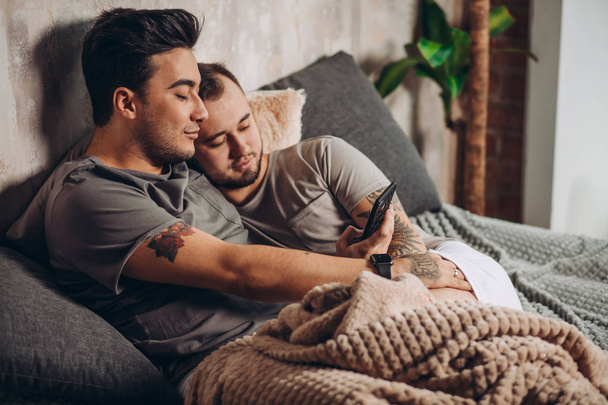 Happy gay couple lying together in bed and using smartphone, looking at photos - Foto, Imagen