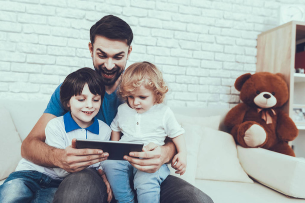 Man Spends Time with His Sons. Father of Boys is Engaged in Raising Children. Father and Sons is Watching a Video on Tablet PC. Persons is Sitting on Couch. People is Located in Bedroom. - Photo, Image