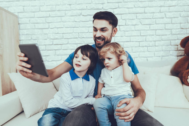 Man Spends Time with His Sons. Father of Boys is Engaged in Raising Children. Father and Sons is Watching a Video on Tablet PC. Persons is Sitting on Couch. People is Located in Bedroom. - Foto, afbeelding