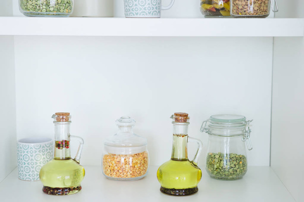 Jars of cereals in the kitchen. Uncooked cereals, grains, beans, olive oil in glass jars on white kitchen shelves. - Фото, изображение