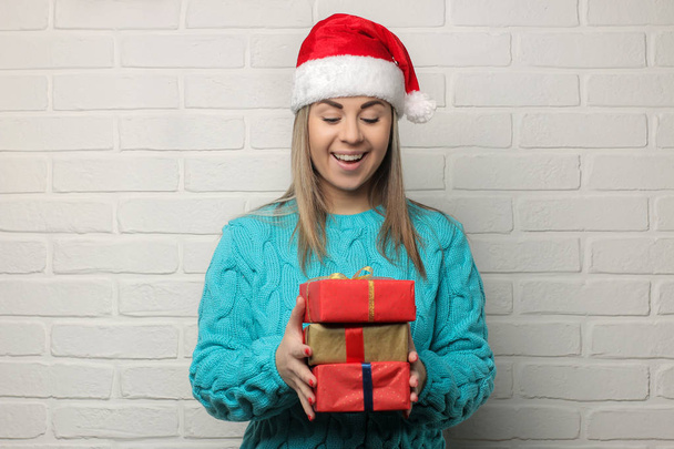 Emotional girl in a Santa Claus hat with Christmas gifts in their hands on a light background - Fotó, kép