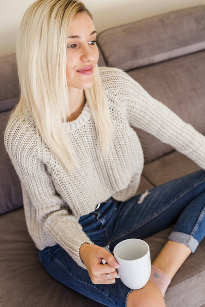 high angle view of happy young woman in jeans and sweater relaxing on couch with cup of coffee - Photo, image