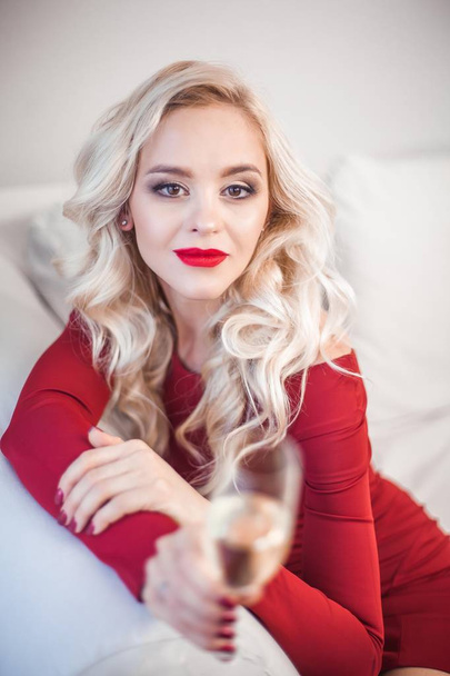 Portret van een mooie jonge blonde met rode lippenstift in een rode jurk. Een jonge vrouw is het bezit van een glas champagne. Vakantie. Kerstmis. Nieuwjaar. - Foto, afbeelding