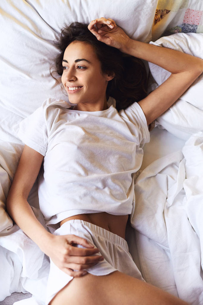 top view of a bright sexual smiling Girl in white t-shirt and shorts, lying on the bed with white bedsheets - Fotoğraf, Görsel