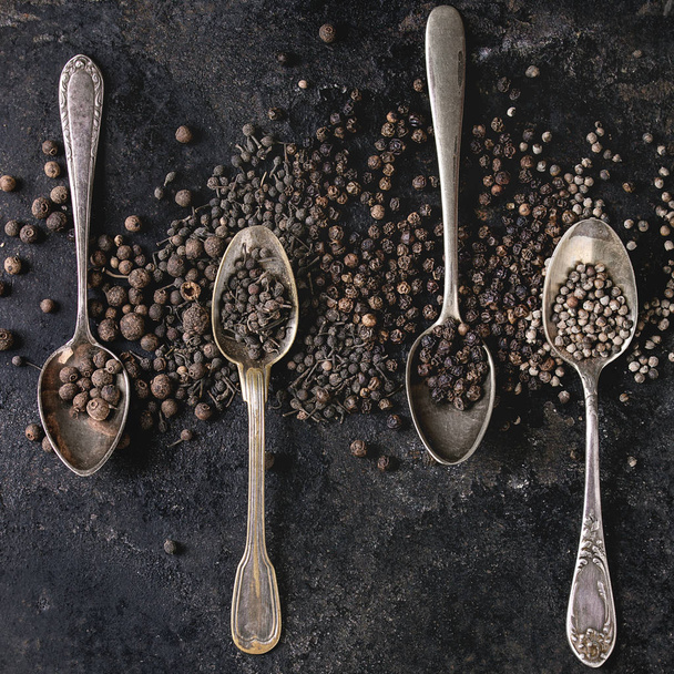 Variety of different black peppers allspice, pimento, monks pepper, peppercorns in vintage spoons over old black iron texture background. Top view, space. Square image - Zdjęcie, obraz