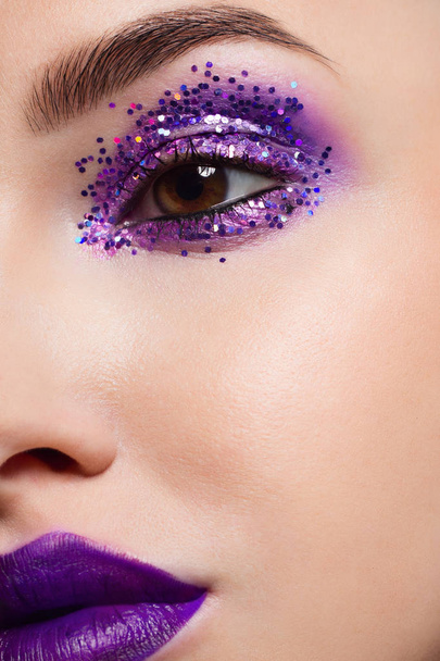 Close-up of the beauty of half a woman's face with creative fashionable make-up of sparkles. Black eyeliner and long eyelashes decorate hazel eyes and lips to match. Well-groomed skin after spa - Фото, изображение