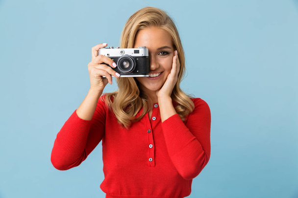 Portrait of cute blond woman 20s wearing red shirt holding retro camera isolated over blue background in studio - Photo, image