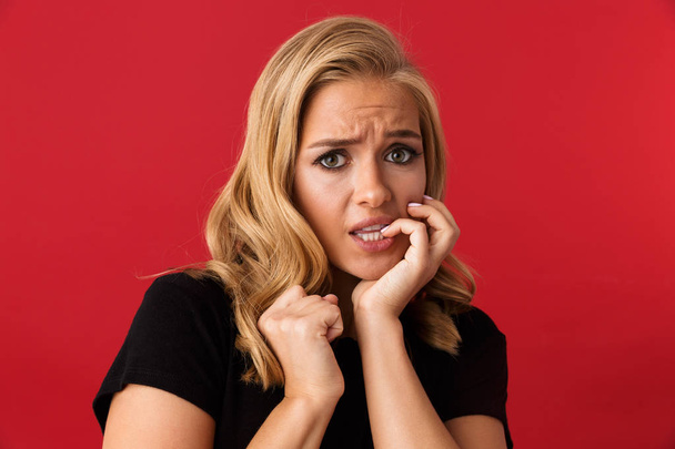 Portrait of frightened blond woman 20s frowning and biting nails isolated over red background in studio - Fotoğraf, Görsel