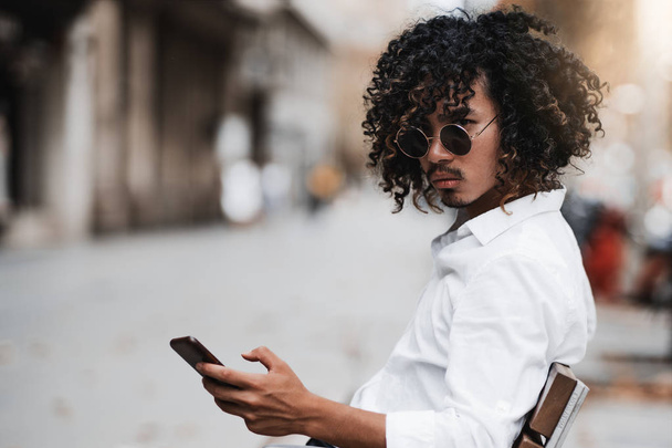 A serious stylish Asian guy with curly hair and in sunglasses is sitting on the bench near a sidewalk and looking aside while typing a message via his smartphone, with a copy space area on the left - Foto, Bild
