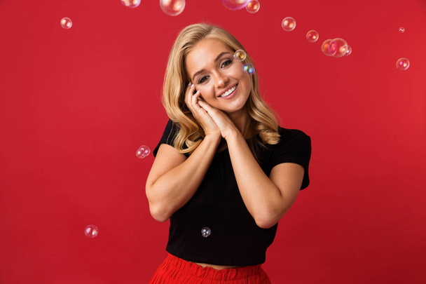 Portrait of gorgeous blond woman 20s smiling at camera with flying bubbles isolated over red background in studio - Zdjęcie, obraz