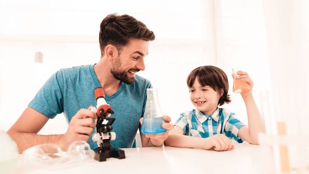 Youn Bearded Father Teaching Son in Shirt at Home. Education at Home. Using Microscope. Studding Chemistry. White Table in Room. Sitting Boy. Young Father. Lesson at Home. Education Concept. - Photo, image