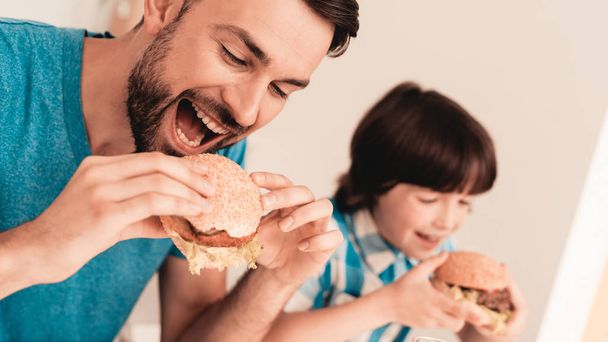 Smiling Son and Father Have Lunch in Kitchen. Boy in Shirt. Modern Kitchen. Sitting Boy. Boy with Spoon. Breakfast in Morning. White Table in Kitchen. Burger in Hands. Young Father. - 写真・画像