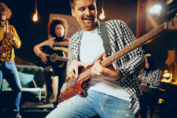 Close up of bass guitarist playing guitar while sitting on the chair. In background rest of the band. Home studio interior. - Photo, Image