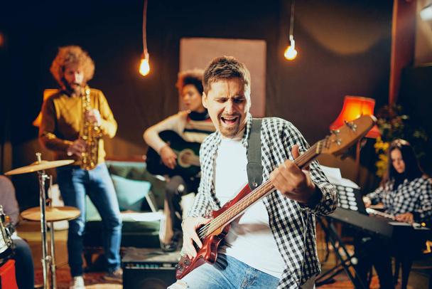 Человек поет и играет на бас-гитаре, сидя в домашней студии. На заднем плане его группа играет на инструментах
. - Фото, изображение