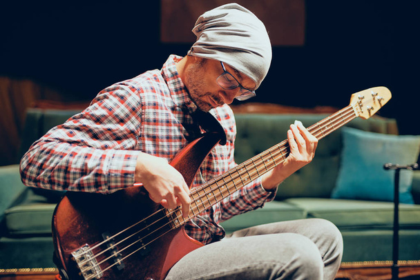 junger kaukasischer Mann mit Hut auf dem Kopf spielt Akustikgitarre, während er im heimischen Studio sitzt. - Foto, Bild