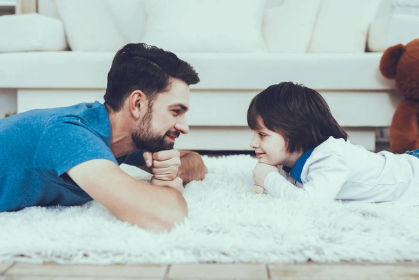 Man Spends Time with His Son. Father of Boy is Engaged in Raising Child. Father and Son is Looking on Each Other. Persons is Lying on Carpet and Smiling. People is Located in Bedroom. - Fotoğraf, Görsel