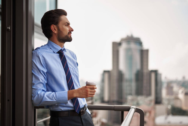 Corporate time-out culture. Waist up side on portrait of businessman leaning doorway of office balcony with cup of coffee and looking at city view - Φωτογραφία, εικόνα