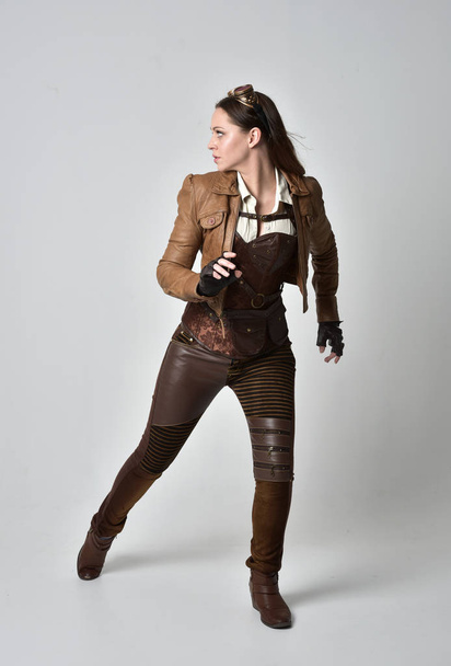 full length portrait of brunette  girl wearing brown leather steampunk outfit. standing pose on grey studio background. - Photo, Image
