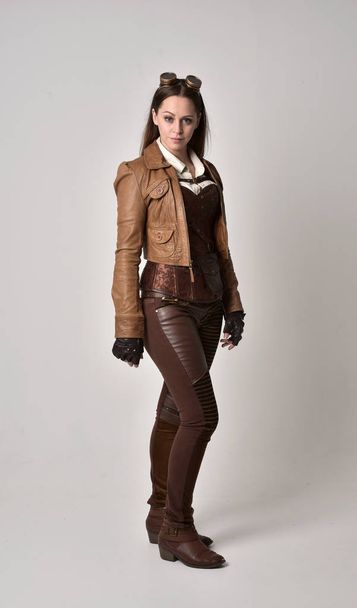 portrait complet de fille brune portant une tenue steampunk en cuir marron. pose debout sur fond de studio gris
. - Photo, image