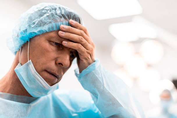 Tired sad doctor frowning and thoughtfully looking into the distance while touching his forehead and having medical mask on the chin - Foto, imagen