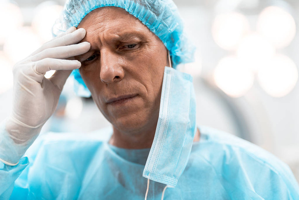 Serious experienced doctor standing alone and touching his forehead with fingers in gloves and thinking - Photo, image